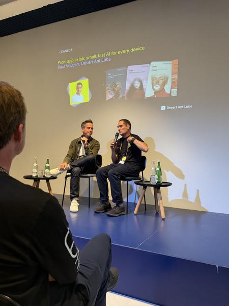 This photo captures an indoor presentation at Groenburgwal, Centrum, Amsterdam, where two men are speaking on a stage with microphones, facing a large projected screen displaying text about AI